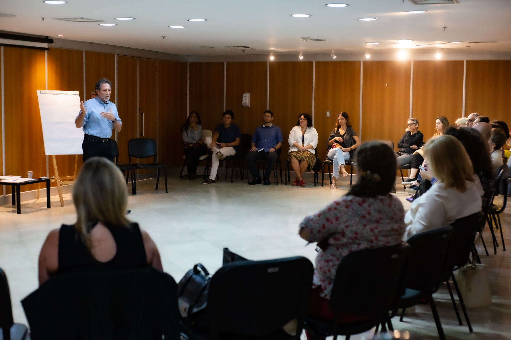 Imagem realizada durante palestra. Ao fundo, palestrante de pé, branco, camisa azul e calça escuro. Atrás dele um quadro branco. Em volta, uma roda formada por mediadores do TJRJ. 