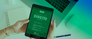 Imagem de duas mãos segurando um tablet, com o texto em tela: Revista de Direito, volume 123. Ao fundo um notebook, um mouse, um caderno e uma caneta.